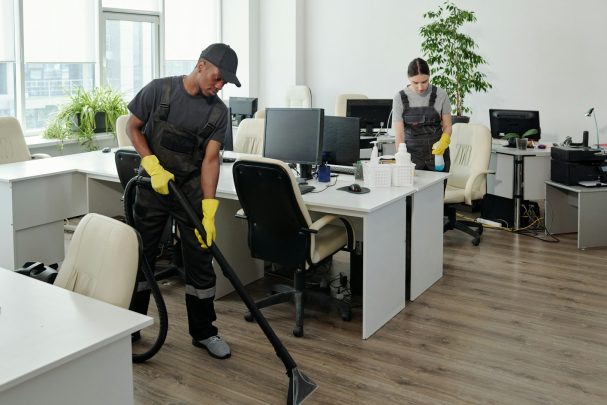 Deux personnes nettoient un bureau avec un aspirateur, portant des gants jaunes.